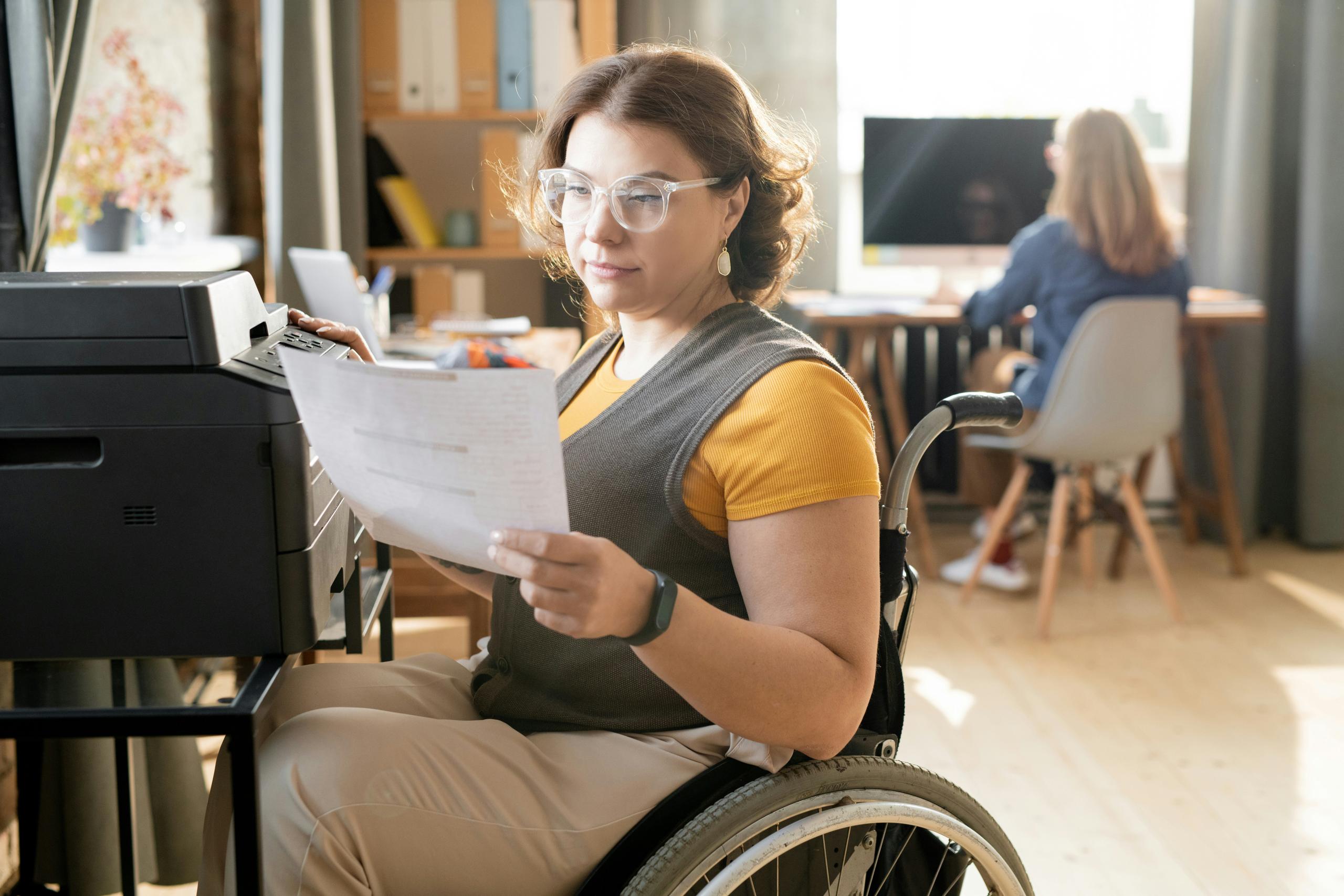 Woman using a wheelchair sitting beside an office printer, holding and reviewing a printed document while wearing glasses and casual work attire, with a modern office environment in the background where another person works at a computer desk.