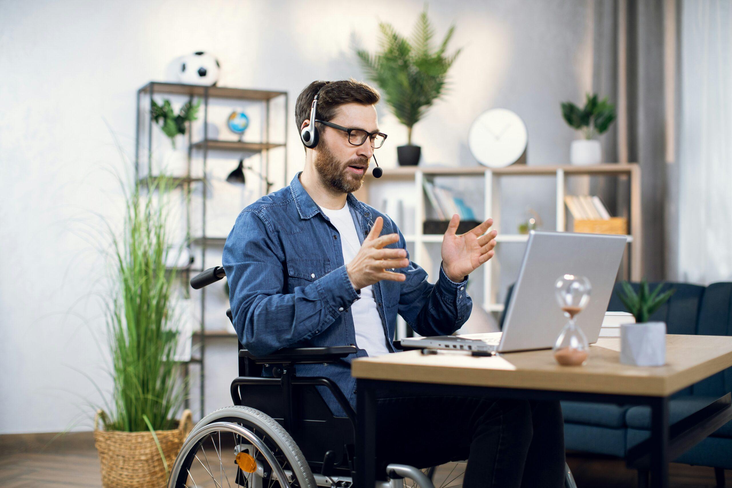 Man in a wheelchair working at a desk with a laptop, wearing a headset and gesturing as if speaking during a virtual meeting in a home office setting