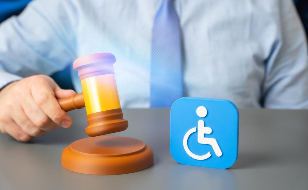 Hand holding a gavel next to a handicap sign on a wooden table, symbolizing justice and accessibility.